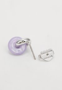 Silberne und lavendelfarbene Ohrstecker im Knopf-Design, einer zeigt einen herzförmigen Rücken mit der Gravur "kate spade new york".