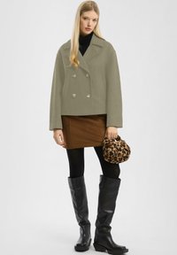 Olijfgroene dubbelbreasted jas, een bruine rok, zwarte panty’s, knie-hoge zwartelaarzen en een handtas met luipaardprint. Algehele eigentijdse look.