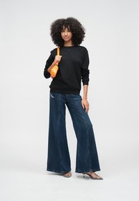 Μαύρο φούτερ, παντελόνι σε στυλ καμπάνα σκούρο μπλε και λαμπερά ασημένια πέδιλα με τακούνι· μοντέλο κρατά μια μικρή πορτοκαλί τσάντα, στέκεται σε λευκό φόντο.