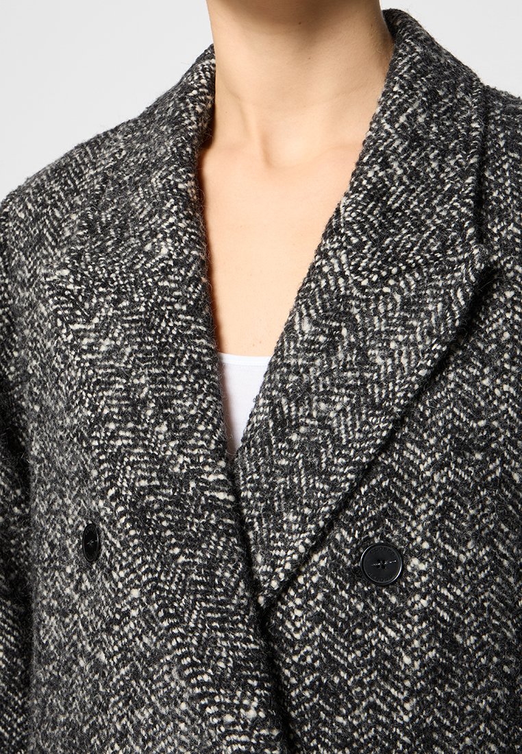 Dubbelgebreide jas in zwart-wit visgraatpatroon. Dikke wolstructuur met een brede kraag en grote zwarte knopen.