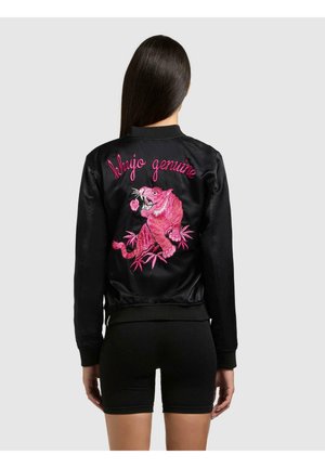 Kobieta z długimi ciemnymi włosami nosi czarną satynową bomberkę z różowym haftowanym tygrysem i napisem "khujo genuine" na plecach.