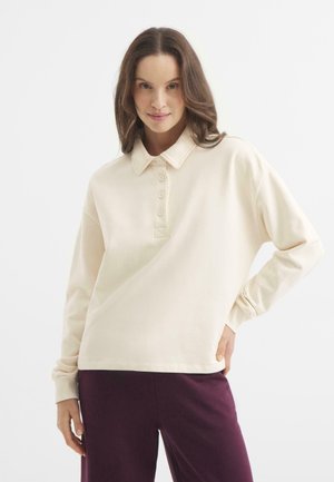 Camisa polo de manga larga color crema con una textura suave, con una tapeta de botones y un corte relajado, combinada con pantalones púrpuras oscuros.