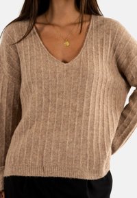 Pull à col en V, en maille côtière, de couleur beige clair, en tissu doux et texturé. Il possède des manches longues et une coupe ample avec un ourlet droit.
