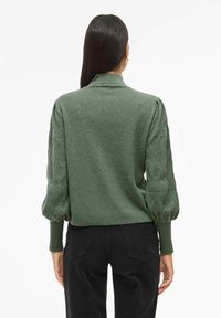 Maglione verde lavorato a maglia con collo alto, maniche a sbuffo e un motivo testurizzato, con un corpo aderente. Pantaloni neri mostrati di seguito.