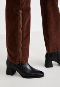 Brown corduroy pants with side zippers worn with black square-toe block-heeled ankle boots on a light background.
