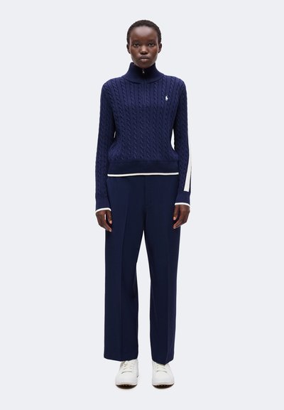 Personne portant un pull torsadé bleu marine avec des rayures blanches et un logo, un pantalon large bleu marine et des baskets blanches, debout devant un fond uni.