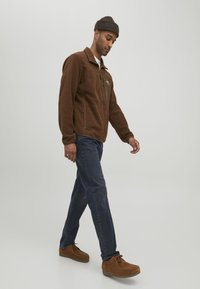 Chaqueta de forro polar marrón con un bolsillo en el pecho con cremallera, combinada con jeans oscuros y zapatos de ante marrones. El modelo lleva un gorro de punto marrón.