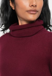 Burgundský rolák vyrobený z mäkkej tkaniny; má preložený golier a hladkú textúru, ktorá zvýrazňuje teplo a pohodlie.