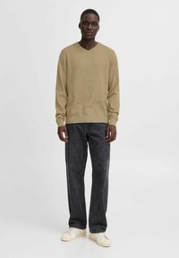 V-hals, langsleeved genser i lys beige strikket materiale, kombinert med mørkegrå jeans og hvite joggesko, satt mot en enkel bakgrunn.