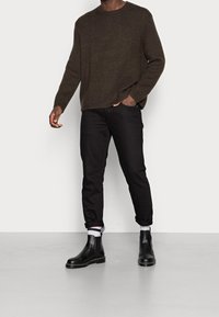 Maglione a maglia marrone scuro, pantaloni neri a sigaretta e stivaletti Chelsea neri. Presenta una texture a coste, polsini arrotolati e accenti di calzini a contrasto.