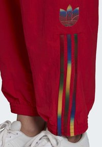 Červené větrovky kalhoty s elastickými manžetami, s barevným pruhovaným detailem po stranách a vyšitým logem. Pod nimi bílé tenisky.