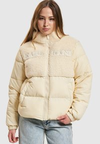 Polstjacka i ljusbeige med krage av konstpäls. Har broderad "KANI," dragkedja och elastiska ärmmuddar. Texturerat och mjukt tyg.