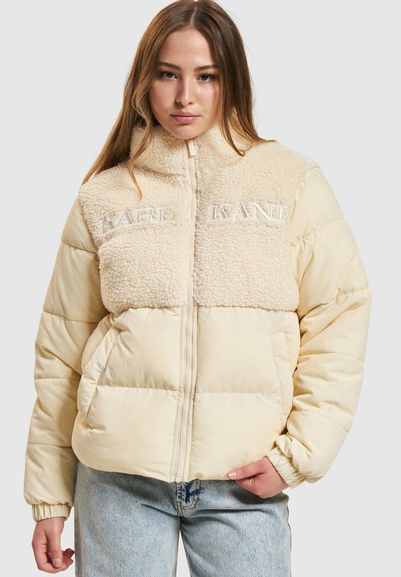 Polstjacka i ljusbeige med krage av konstpäls. Har broderad "KANI," dragkedja och elastiska ärmmuddar. Texturerat och mjukt tyg.