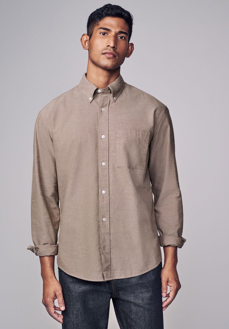 Camicia button-up marrone chiaro con maniche lunghe, taschino sul petto e polsini leggermente arrotolati. Tessuto testurizzato e orlo curvo.