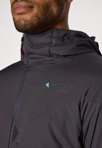 Schwarze, leichte Jacke mit hohem Kragen, Reißverschluss und atmungsaktivem Stoff; verfügt über ein blaues Logo und ein dezentes strukturiertes Muster.