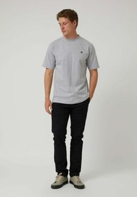 Camiseta gris de manga corta con un cuello redondo y un pequeño logo, combinada con jeans oscuros y calzado claro, de pie frente a un fondo neutro.