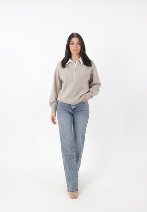 Pull beige avec une chemise blanche à col en dessous, associé à un jean droit bleu clair. Le modèle se tient sur un fond blanc.