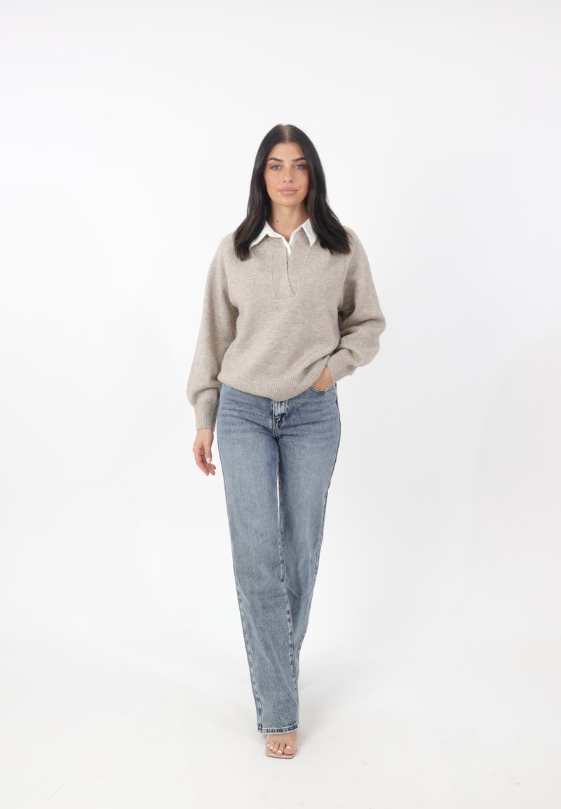 Maglione beige con una camicia bianca con colletto sotto, abbinato a jeans dritti azzurri chiari. Il modello è in piedi su uno sfondo bianco.