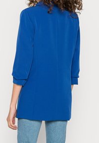 Blazer bleu ajusté avec manches trois-quarts, une texture lisse et un col pointu. Dispose d'une seule couture au dos et aucun élément métallique visible.