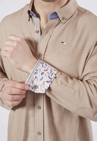 Chemise beige à manches longues avec un emblème brodé, col boutonné et poignets à motifs avec des feuilles de couleurs variées. Boutons décoratifs visibles.