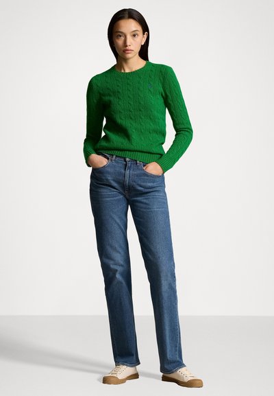 Pull en laine vert en tricot torsadé avec un col rond, associé à un jean bleu droit et des baskets beiges. Le pull présente des poignets et un ourlet côtelés.