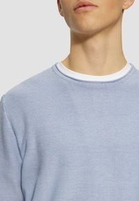 Maglione azzurro chiaro a girocollo realizzato in tessuto testurizzato, con un colletto interno bianco. Vista ravvicinata che mette a fuoco l'area del collo e della spalla.