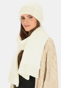 Biały zestaw czapki beanie i szalika, z prążkowaną teksturą i miękkim materiałem. Szalik swobodnie układa się wokół szyi.