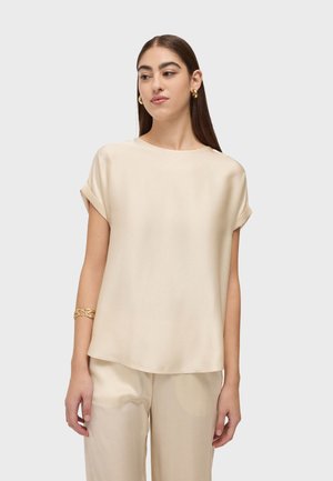 Jeune femme aux longs cheveux bruns portant un chemisier beige à manches courtes et un pantalon assorti, des boucles d'oreilles créoles en or et un bracelet en or.