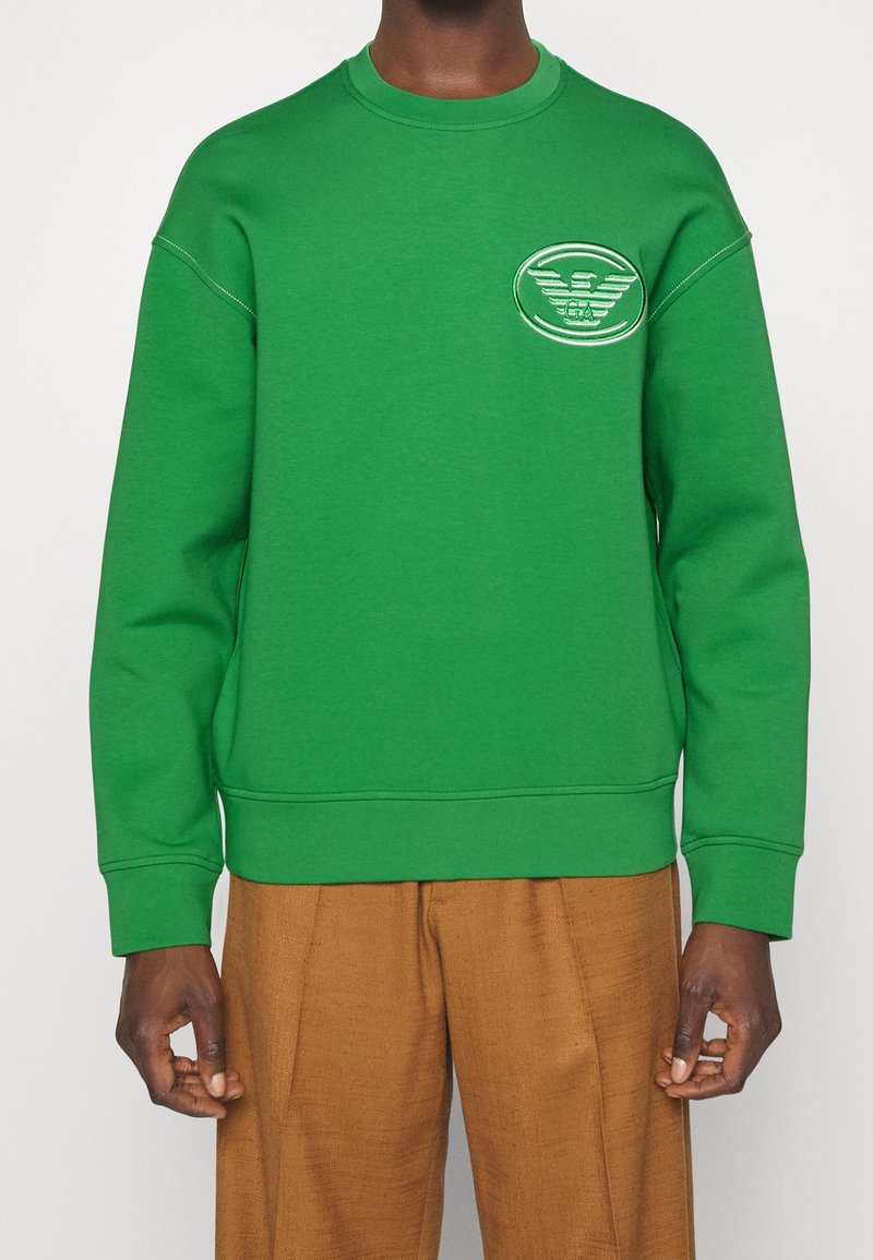 Felpa verde con polsini e orlo a costine, caratterizzata da un logo ricamato sul petto. Indossata con pantaloni tan strutturati.