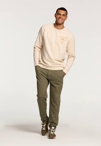 Ljust beige sweatshirt med ett triangulärt mönster på bröstet, kombinerad med mörkgröna joggingbyxor och bruna sneakers med vita detaljer.