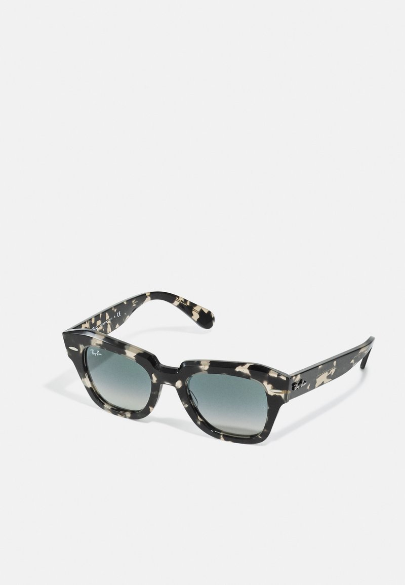 Melna un krēmīgi rakstainas saulesbrilles ar biezu rāmju, kvadrātveida formu un gradienta tonētu stiklu. Ray-Ban logo sānos.