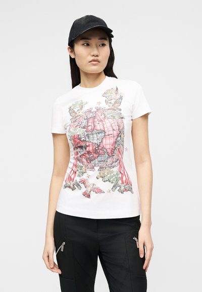 Νεαρή γυναίκα φορώντας μαύρο καπέλο, λευκό μπλουζάκι με σχέδιο κόκκινου και γκρι παγκόσμιου χάρτη και πουλί, και μαύρο παντελόνι με τσέπες με φερμουάρ.