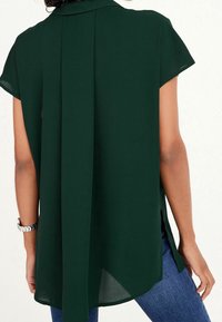 Blouse vert foncé à manches courtes, avec un pli dans le dos et des fentes sur les côtés. Fabriquée en tissu léger, elle comporte un col et un ourlet arrondi.