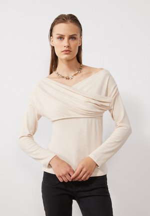 Beige long-sleeve top with a draped, off-shoulder design and gathered fabric. Paired with dark jeans; features a gold statement necklace.