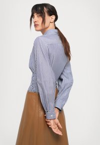 Camisa de rayas azul y blanca de manga larga con un ajuste ceñido en la parte posterior, que presenta puños abotonados y detalles fruncidos en la cintura, combinada con una falda marrón.