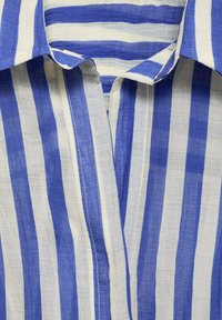Blau-weiß gestreifter Stoff mit einer strukturierten Webart, der einen Kragen und Knöpfe an der Vorderseite aufweist. Die Streifen variieren in der Breite.