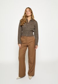 Rjavo pulover z zadrgo in rebrastim zaključkom, kombiniran z lahkimi svetlo rjavimi hlačami širokih nog in bež petami z ostrimi konicami. Preprost, teksturiran dizajn.