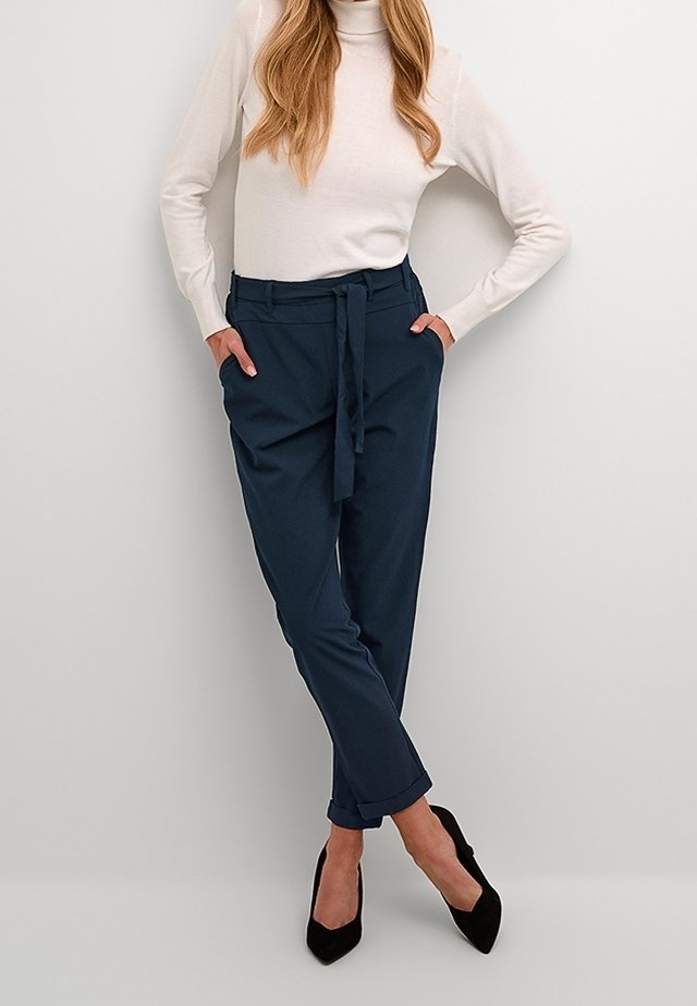 Femme debout, jambes croisées, portant un col roulé blanc, un pantalon bleu marine ceinturé et des talons noirs sur un fond clair uni.