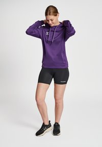Felpa sportiva viola con tasca frontale, cappuccio con cordino e logo. Abbinata a pantaloni corti neri e scarpe da ginnastica nere.