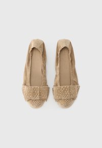 Μπεζ σουέντ φλατ παπούτσια με επίπεδη σόλα, ελαφρώς μαζεμένο πάνω μέρος και φαρδιά φιόγκο με μεταλλικά καρφιά στη μύτη. Ομαλή υφή σε όλο το παπούτσι.