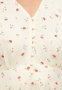 Κρεμ φόρεμα με μικρά κόκκινα και ροζ λουλουδάτα σχέδια, κουμπιά καλυμμένα με ύφασμα μπροστά και λεπτομέρεια με συγκέντρωση στη μέση.