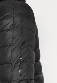 Svart vadderad jacka med quiltad textur. Har en glänsande logotyp längs sidan med en glansig finish och erbjuder isolering och värme.