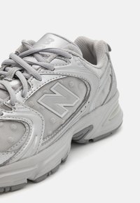 Šedá sportovní teniska s texturovaným síťovaným svrchním dílem, reflexními detaily a výrazným logem „N“. Má tkaničky a polstrovanou podrážku.