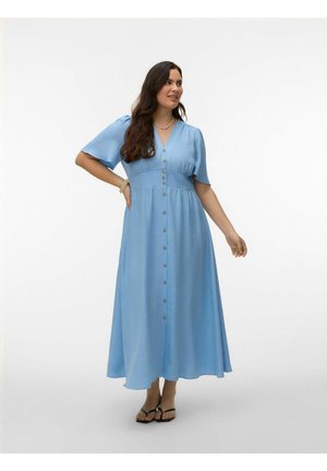 Mujer con pelo largo y oscuro, vestida con un vestido largo azul claro con botones al frente, lleva sandalias negras y joyería sencilla, de pie contra un fondo blanco.