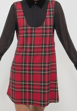 Rotes Tartankleid mit Trägern, getragen über einer schwarzen, durchsichtigen Langarmbluse mit spitzem Kragen und geknöpftem Vorderteil.