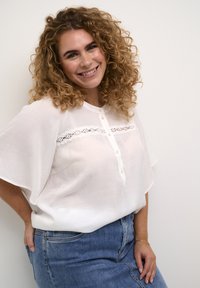 Hvid bluse med korte, løse ærmer og knaplukning, med en blondestykke hen over brystet; parret med et denimskørt.