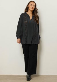 Sort bluse med lange ærmer og V-hals, der har intrikate blonder detaljer langs fronten og ærmerne, kombineret med sorte vidde bukser.