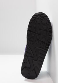 Semelle de baskets Nike en caoutchouc noir avec une adhérence texturée, présentant un motif en zigzag proéminent et un logo. Le produit est appuyé contre une surface blanche.