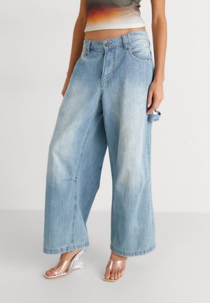 Femme portant un jean denim bleu clair à jambes larges et des sandales compensées transparentes, debout devant un fond uni.
