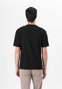 Tricou negru cu mânecă scurtă, cu un design texturat, gât rotund și croială dreaptă, asortat cu pantaloni bej, văzut din spate.
